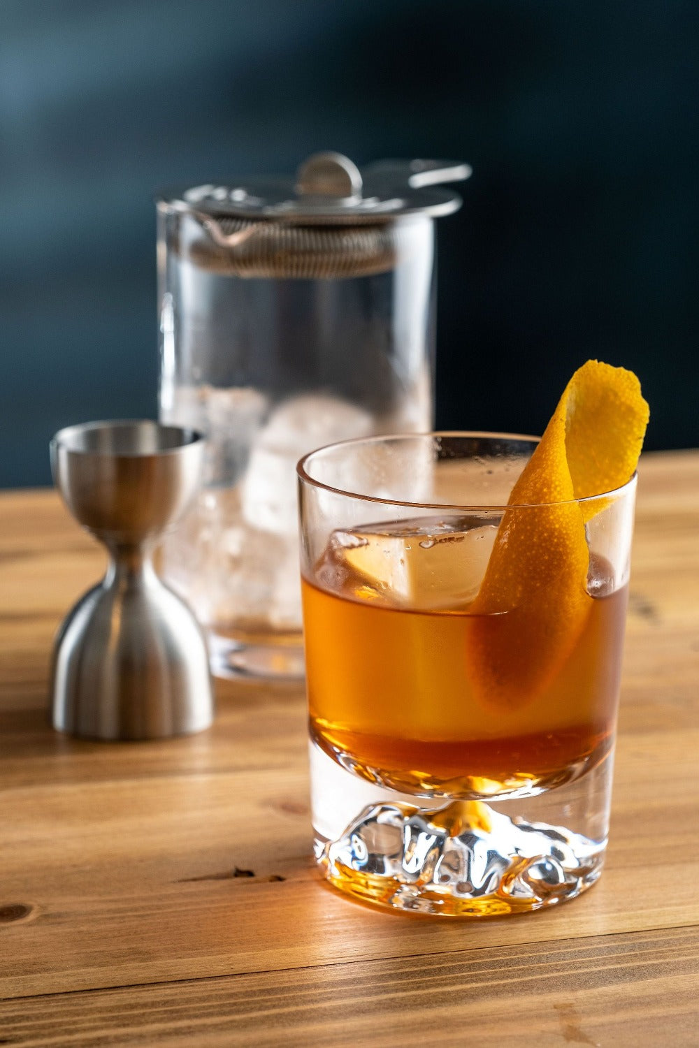 Glass of amber cocktail with an orange twist on a wooden bar with a cocktail shaker in the background.