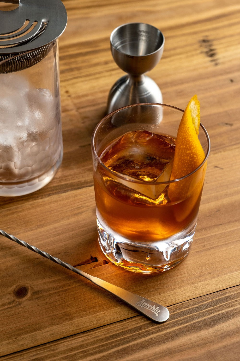 Glass of amber cocktail with an orange twist on a wooden surface, accompanied by a jigger and strainer.