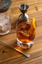 Glass of amber cocktail with an orange twist on a wooden surface, accompanied by a jigger and strainer.