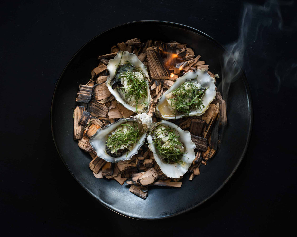 Four oysters on a bed of wood with smoke in a dark setting