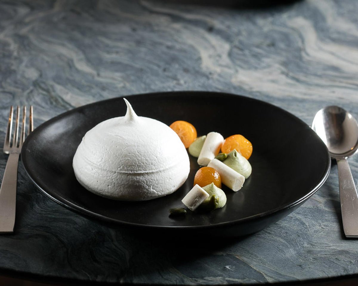White dessert on a black plate with oranges and white cubes, served on a marble surface.