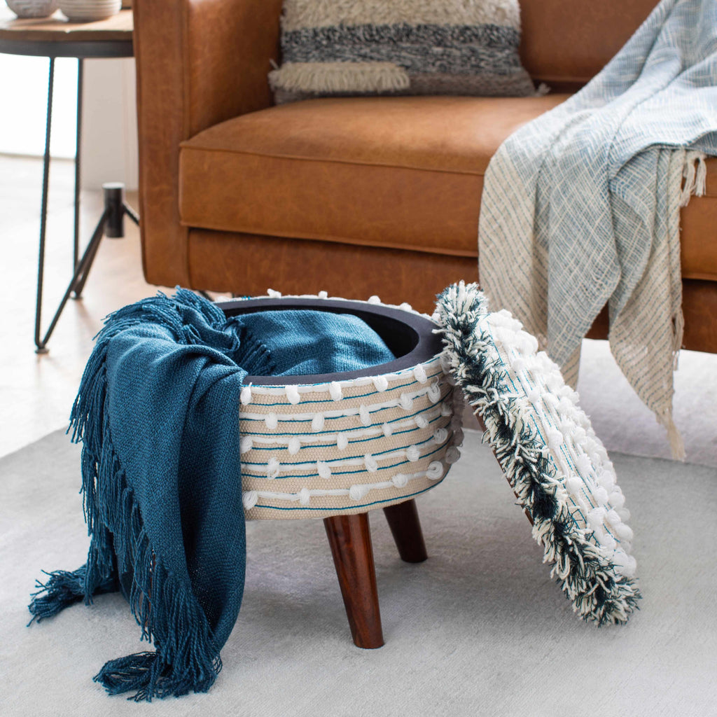 Decorative footstool with a woven design and wooden legs, draped with blue and white textured cloths, in a living room setting.