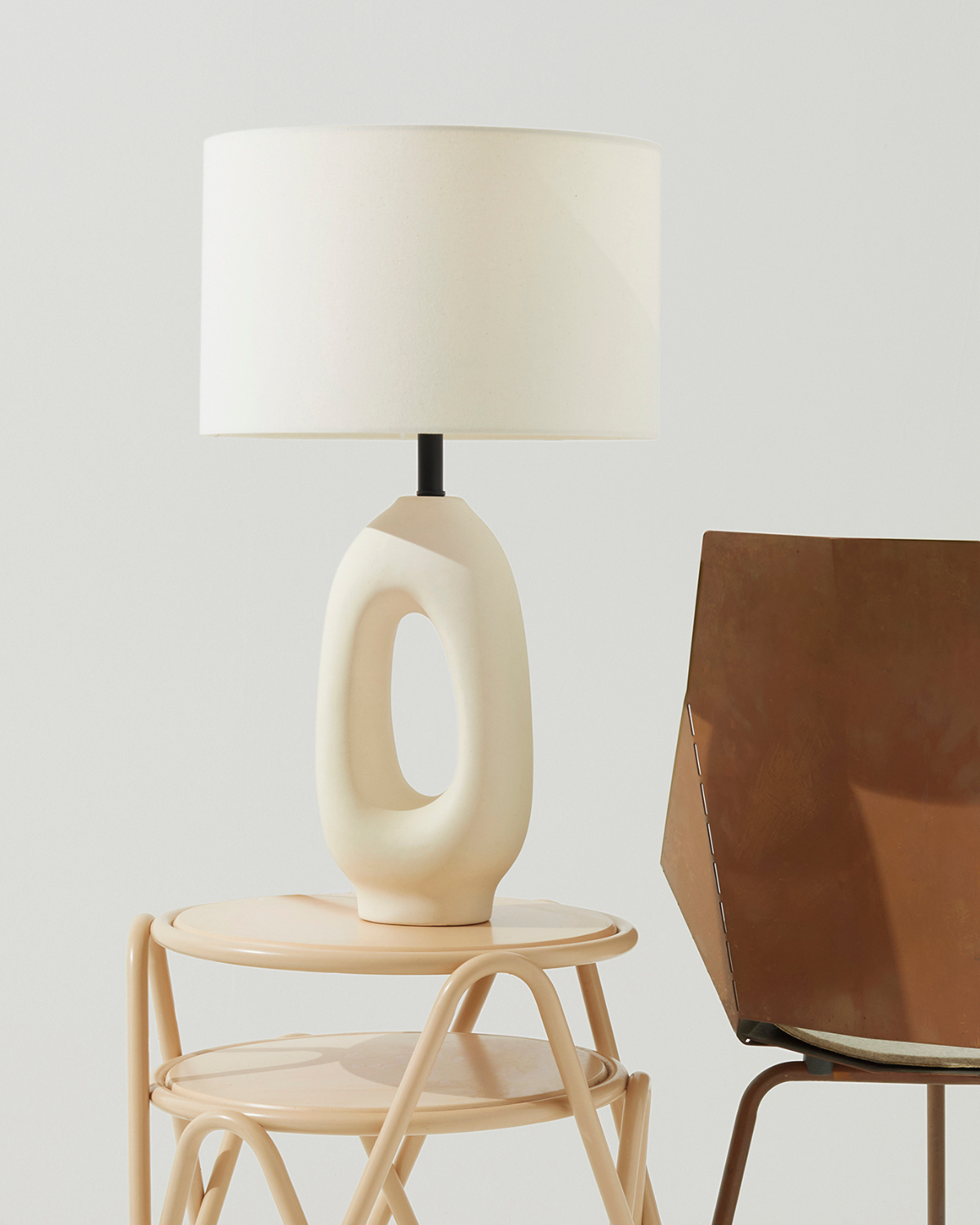 An off-white sculptural table lamp with a cylindrical shade and looped base, displayed on a light wood side table next to a brown leather chair against a neutral light gray background.