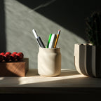 Ceramic pen holder with pens on a wooden surface with a plant and wooden box in the background.
