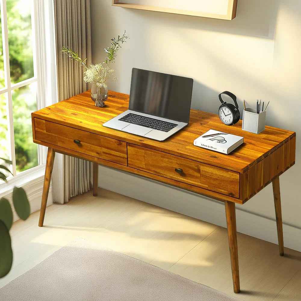 Wooden desk with laptop, clock, and stationery in a bright room.
