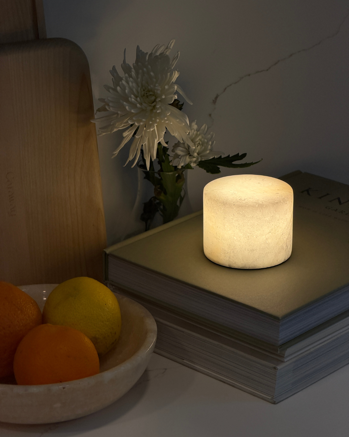 Small lit candle like lamp on a stack of books with a bowl of oranges and flowers in the background.
