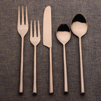 Set of rose gold cutlery including forks, knife, and spoons on a textured brown background.