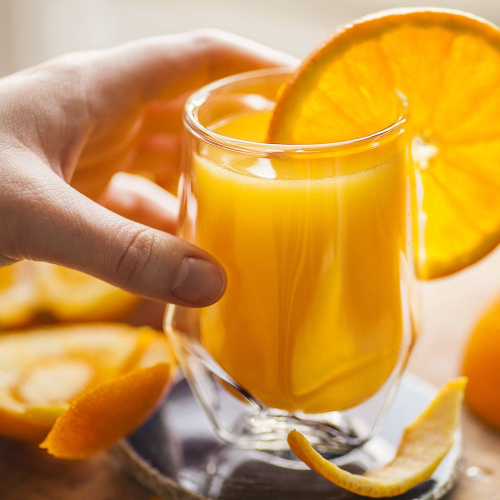 Glass of orange juice with a slice of orange, surrounded by more oranges on a wooden surface.