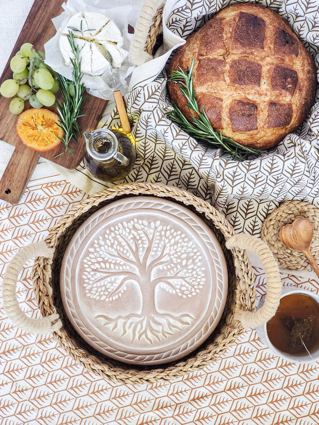 Decorative plate with tree design on a woven basket, surrounded by bread, fruit, and herbs on a patterned tablecloth.