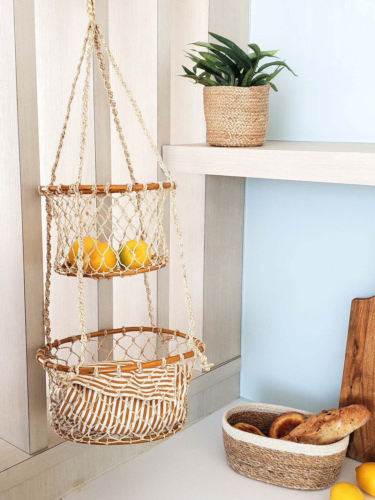 Hanging macrame basket with fruits on a shelf with a plant and bread basket in the background.