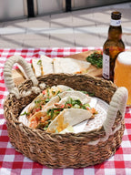 Wicker basket with tacos on a red and white checkered tablecloth, accompanied by a bottle and glass of beer.