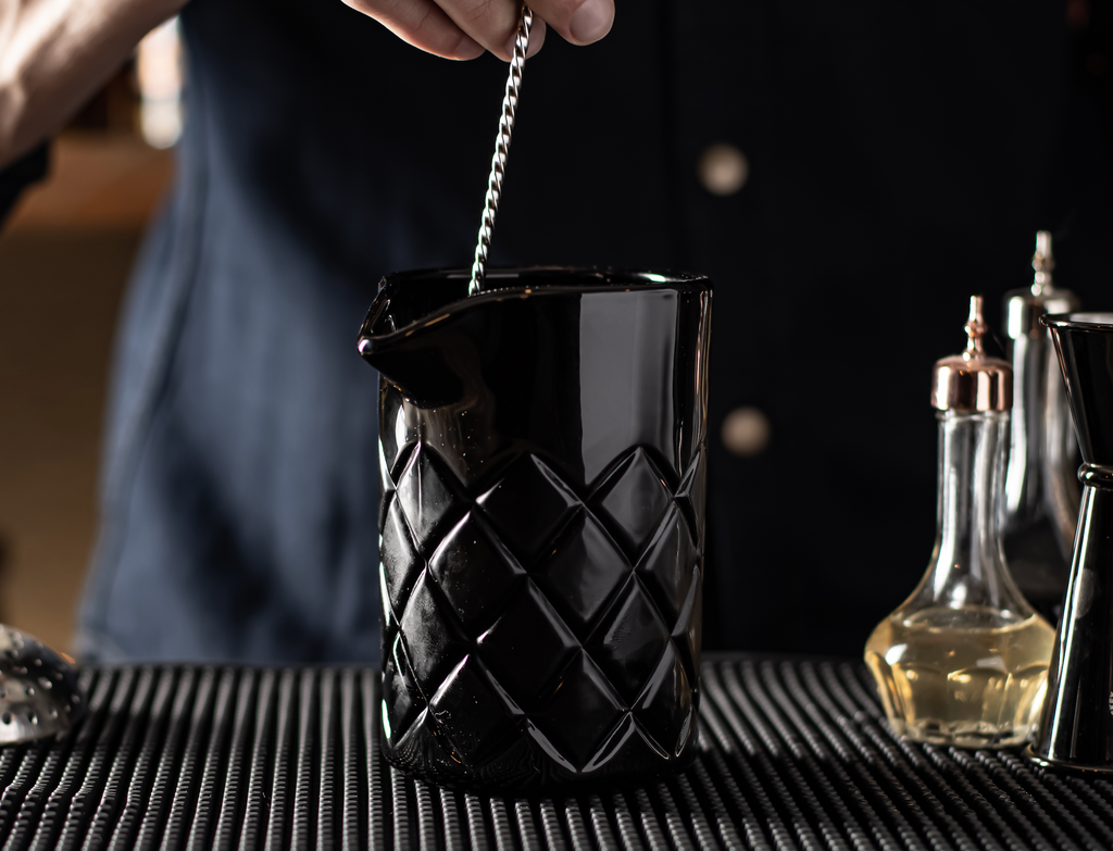 Person stirring a cocktail in a black glass with a textured pattern on a bar.