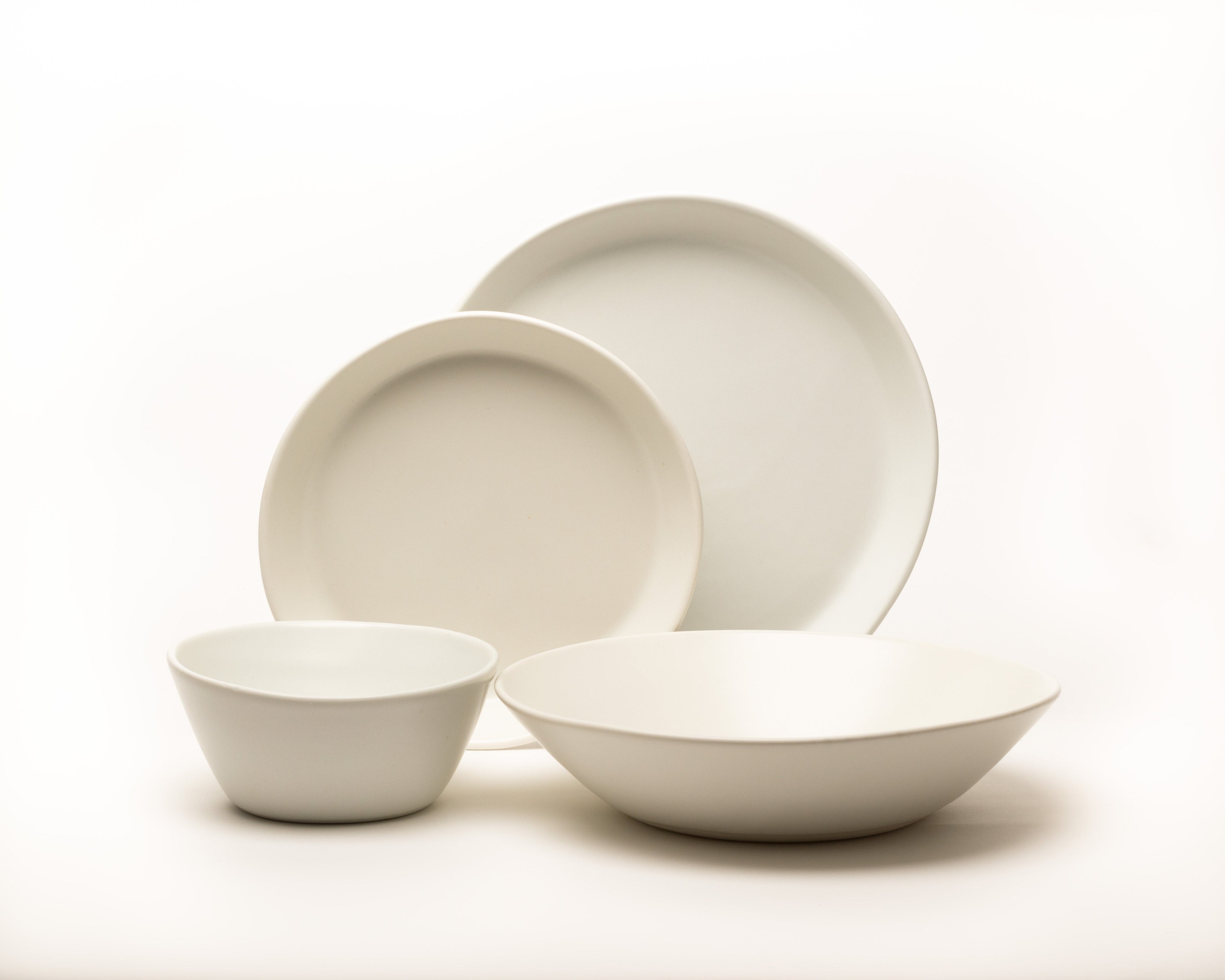 Set of white ceramic dishes including a bowl, two plates, and a cup on a light background