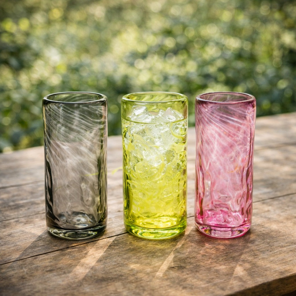 Three colorful glass tumblers on a wooden surface with a blurred green outdoor background.