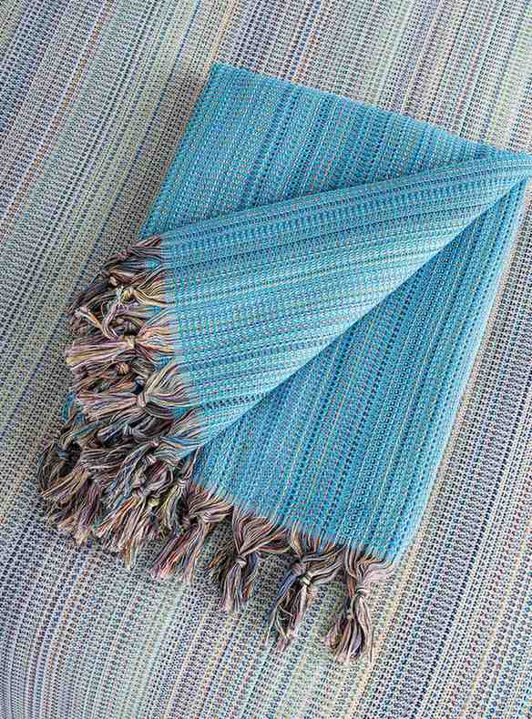 Folded turquoise king size blanket with multicolor tassels on a textured surface