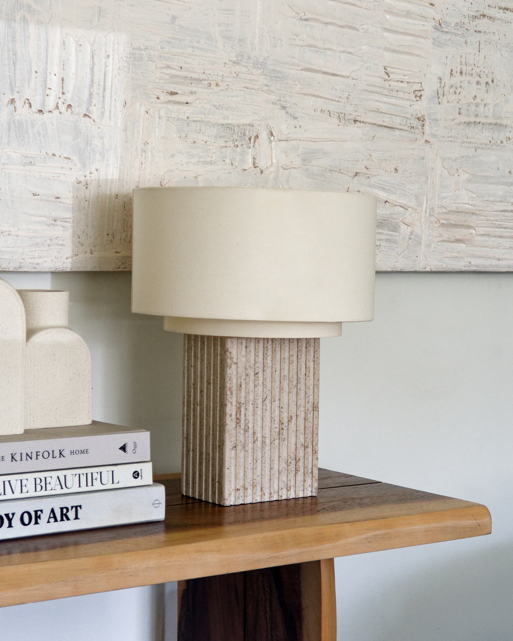 Beige table lamp with textured base on a wooden surface against a textured wall.