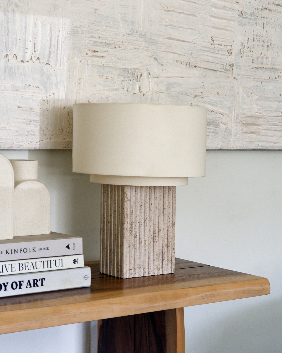 Beige table lamp with textured base on a wooden surface against a textured wall.