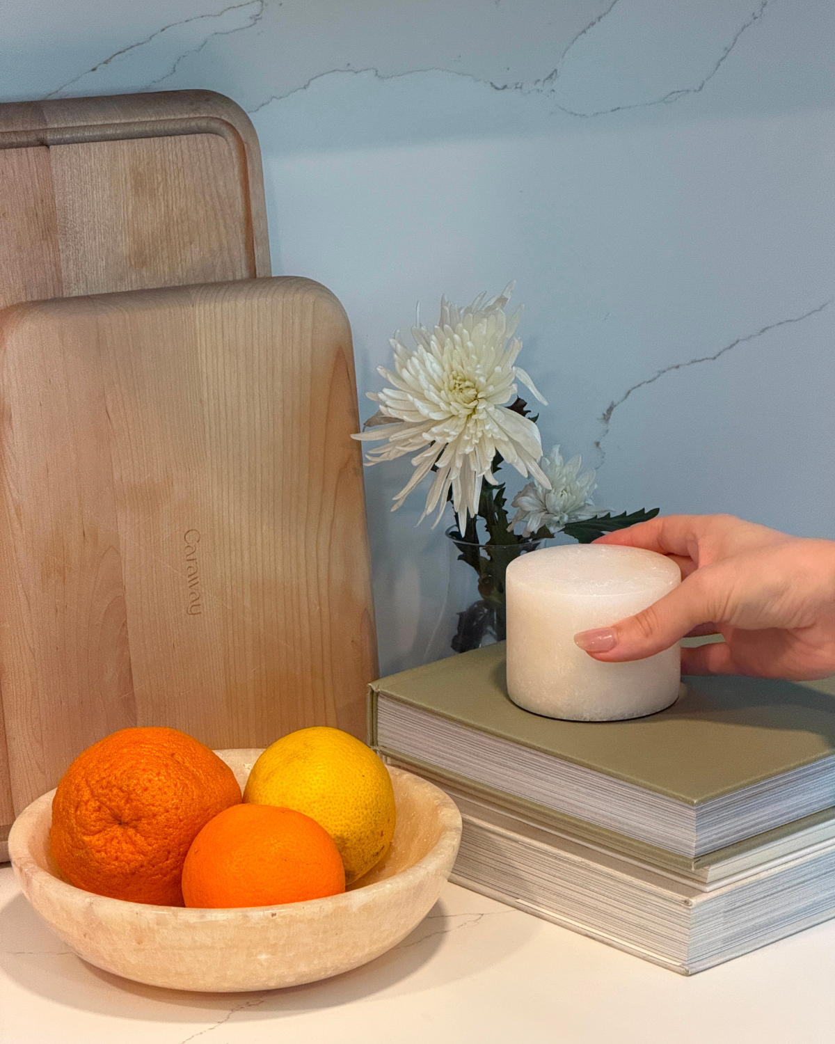Bowl of oranges, wooden cutting board, and a hand holding a candle like lamp on a light blue background.