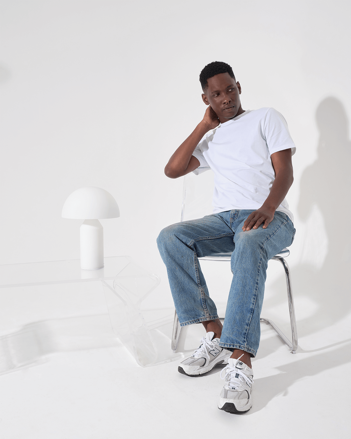 Person sitting on a chair wearing a white t-shirt, blue jeans, and white sneakers in a minimalistic setting.