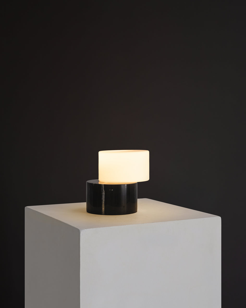 Modern table lamp with a black base and white shade on a gray pedestal against a dark background