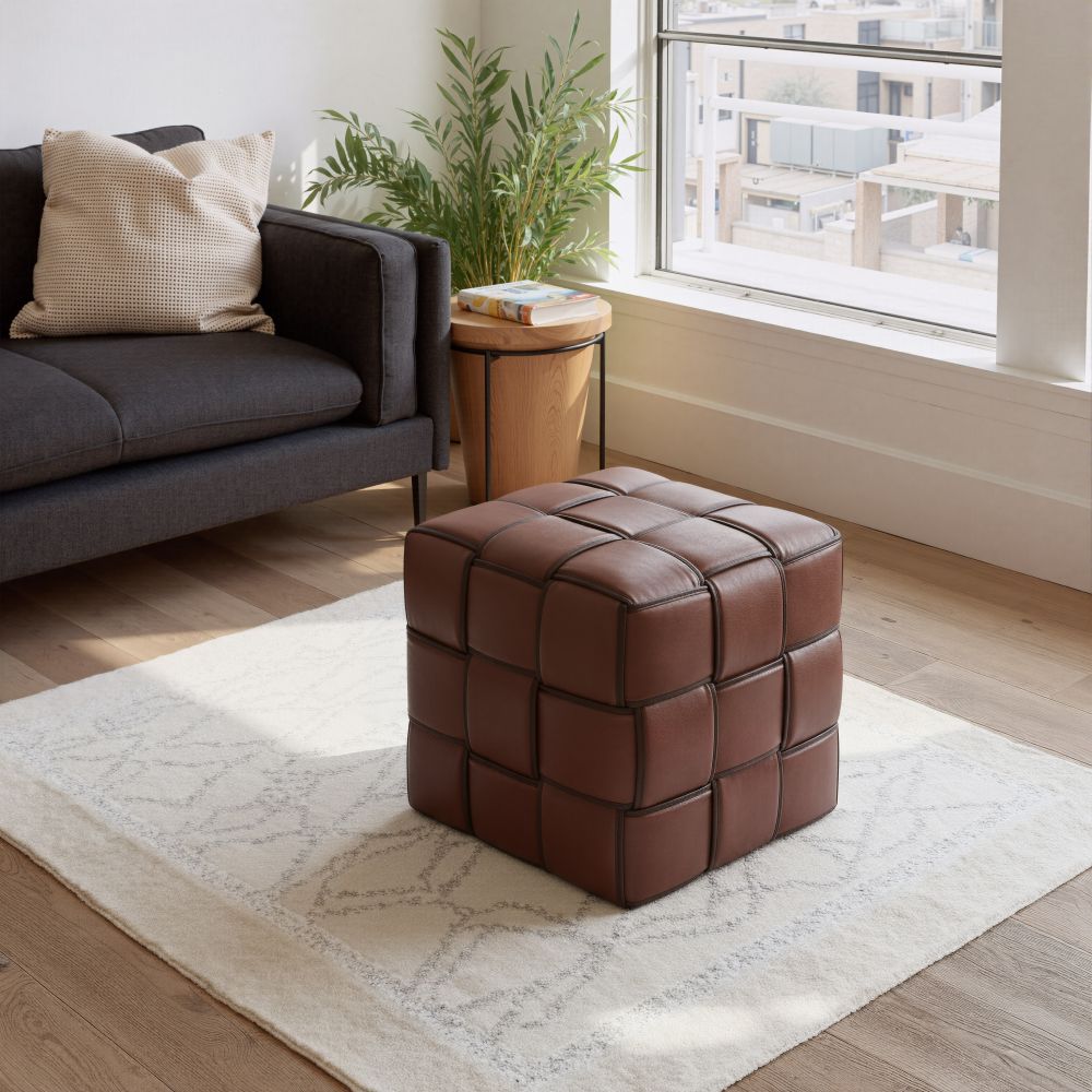 Brown leather cube ottoman in a living room setting with a sofa and window in the background.