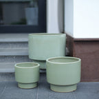 Three light sage green ceramic planters in nested sizes placed on a dark tiled floor next to a doorway.