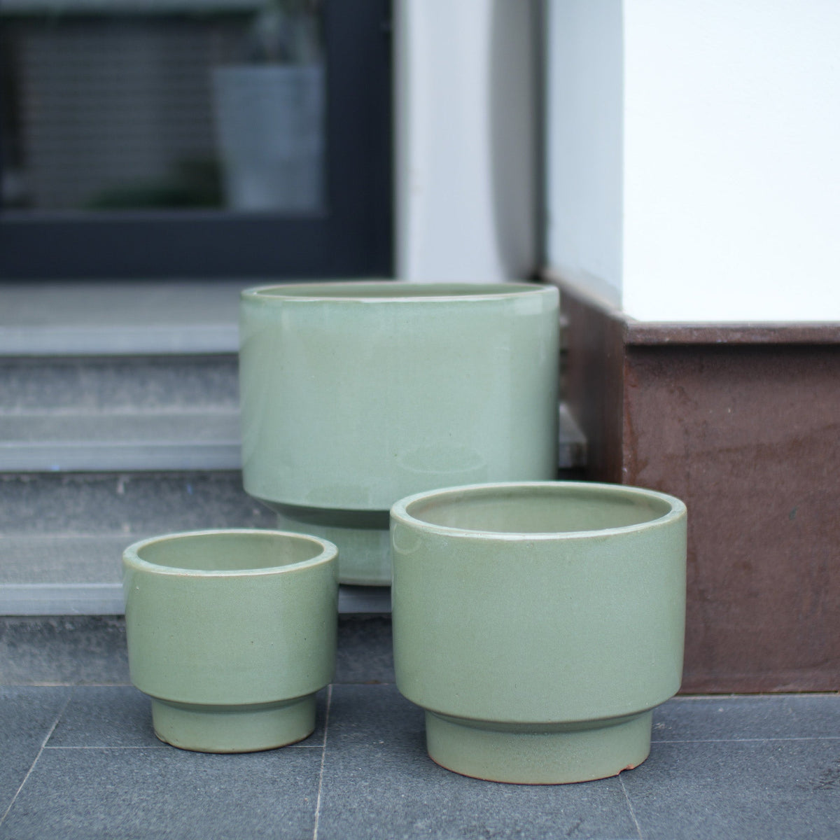Three light sage green ceramic planters in nested sizes placed on a dark tiled floor next to a doorway.