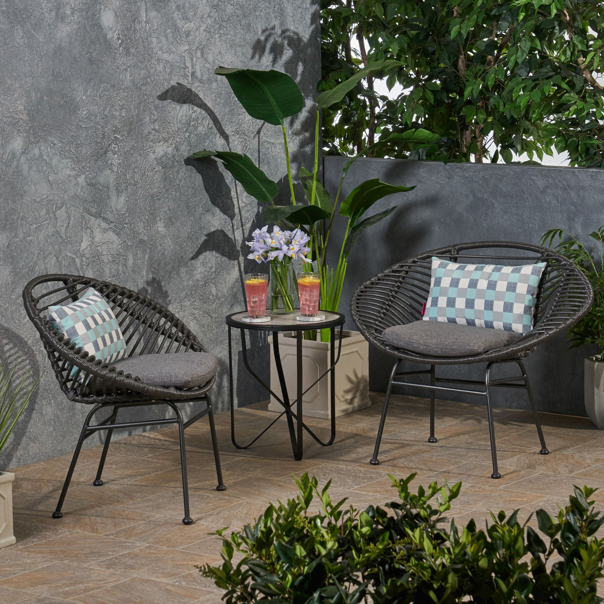 Outdoor patio set with two chairs, a small table, and decorative pillows on a patio with plants and a stone wall.