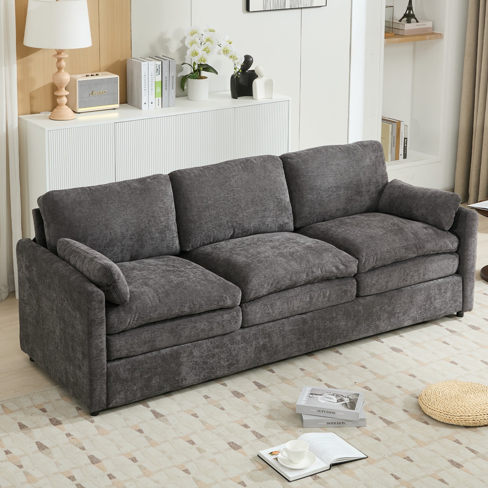 A three-seat gray fabric sofa in a modern living room setting, positioned in front of a white sideboard with decorative items, books, and a lamp.