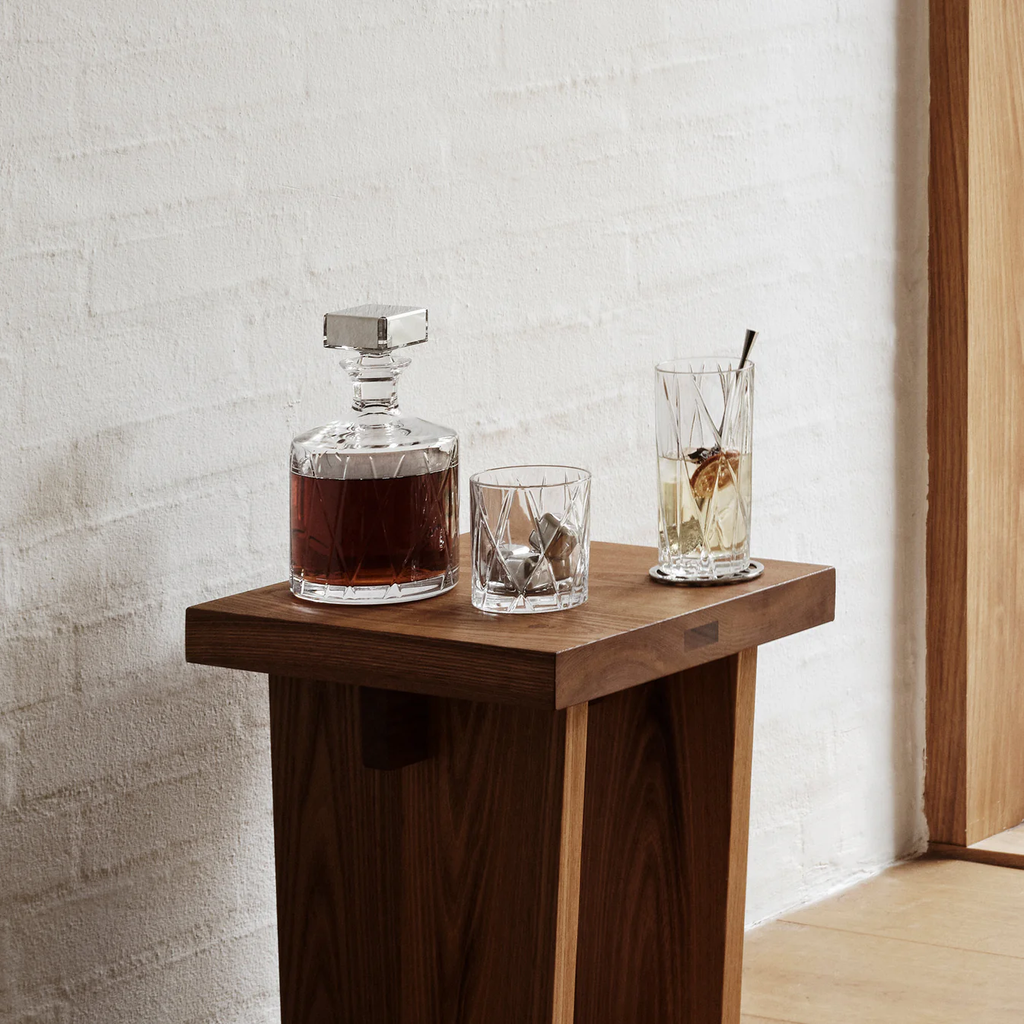 Wooden side table with a decanter and glasses against a light brick wall.