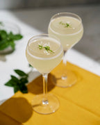 Two glasses of a light-colored cocktail with garnishes on a yellow cloth.