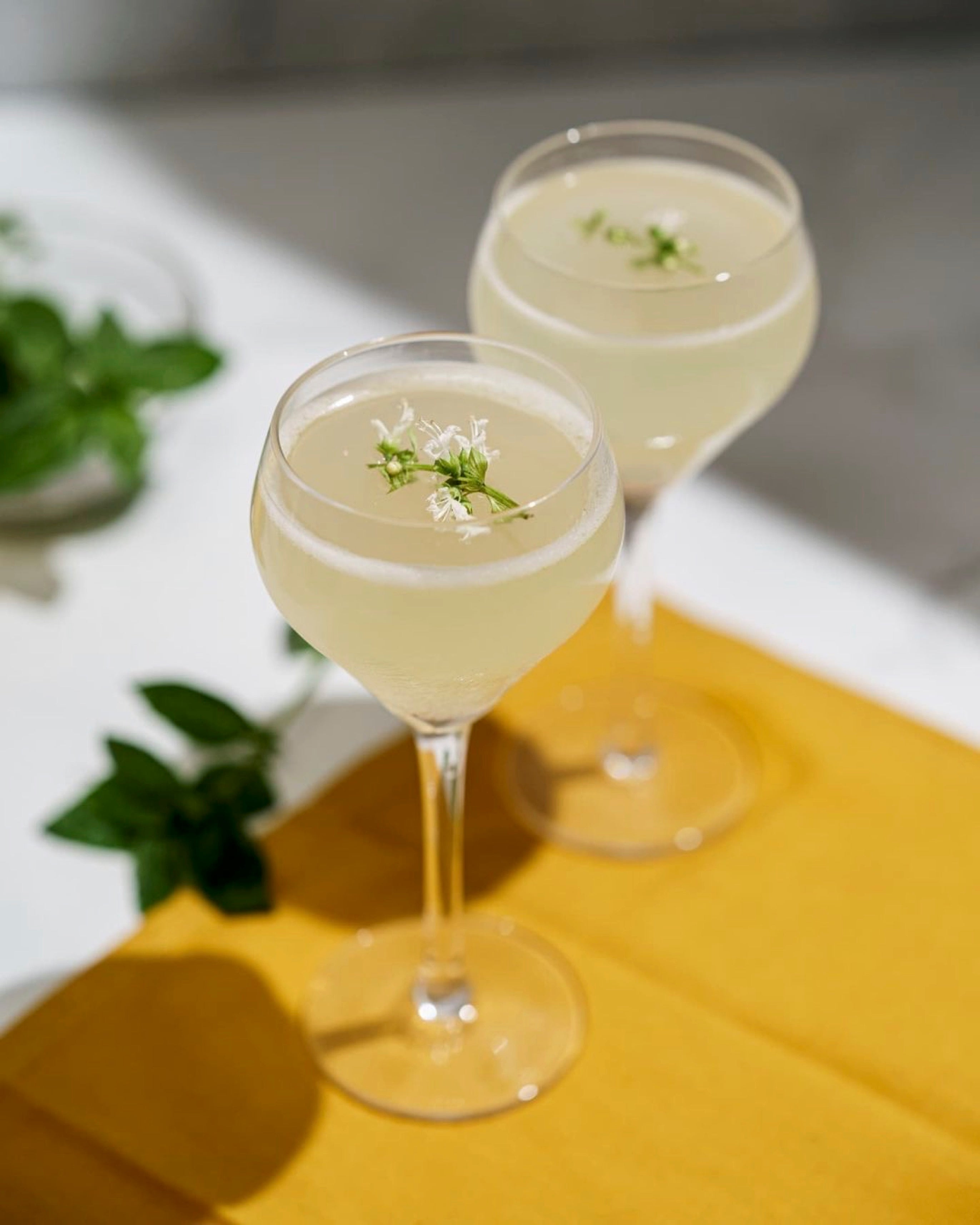 Two glasses of a light-colored cocktail with garnishes on a yellow cloth.