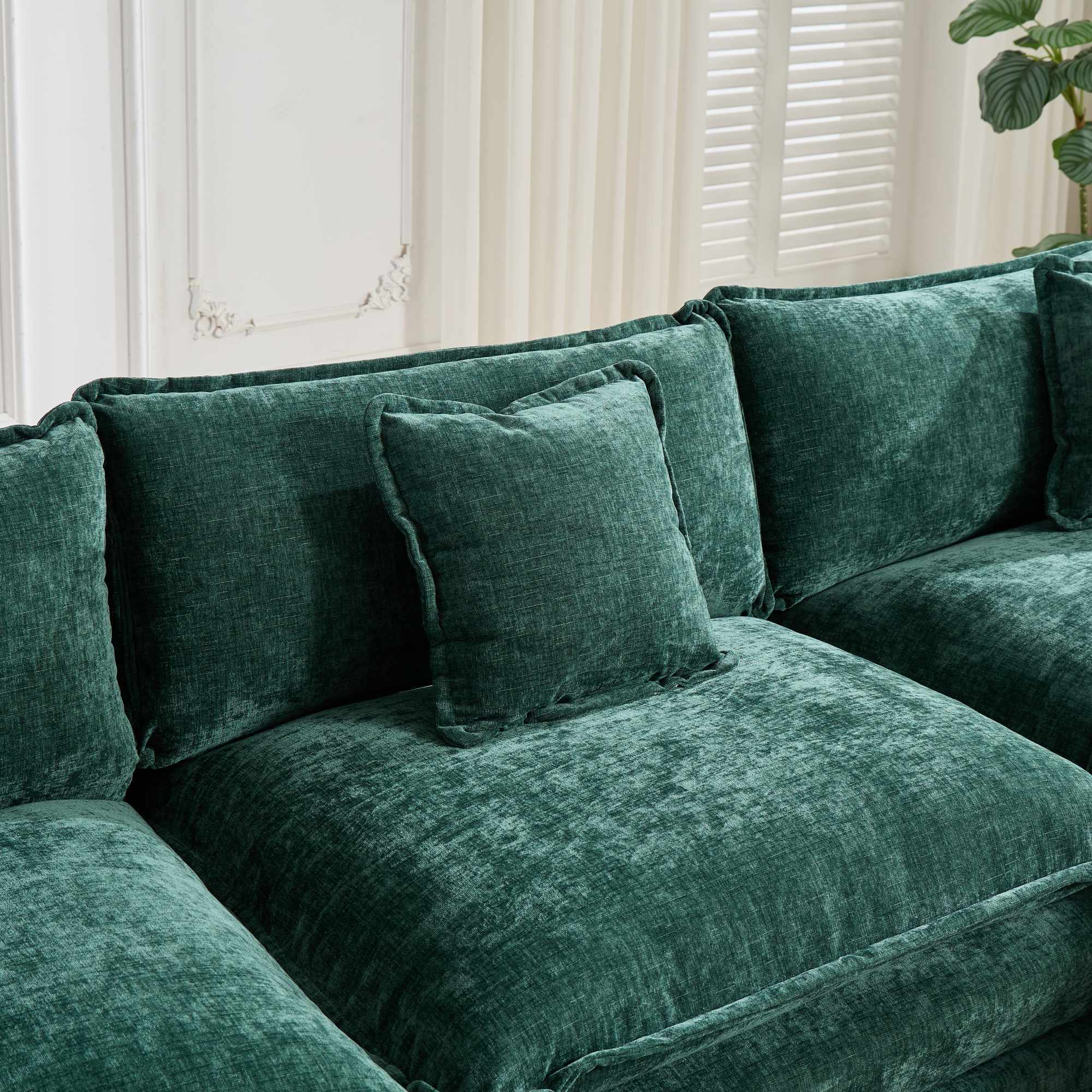 Green velvet sectional sofa with cushions in a room with white walls and a plant.