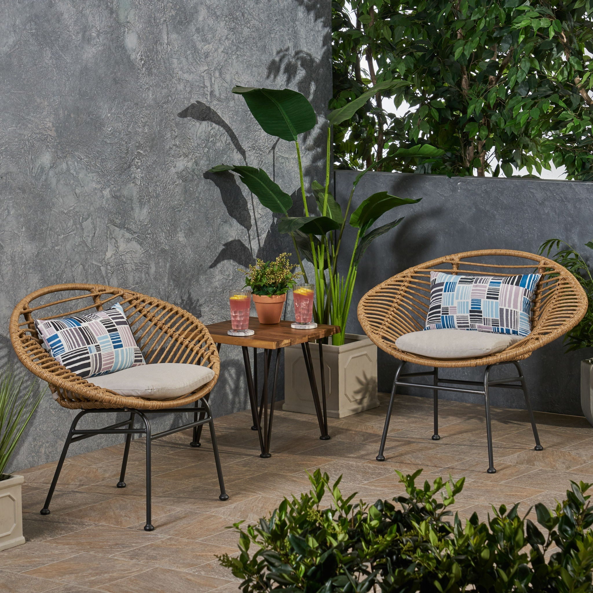 Outdoor patio set with two wicker chairs, a small table, and decorative pillows against a gray wall.