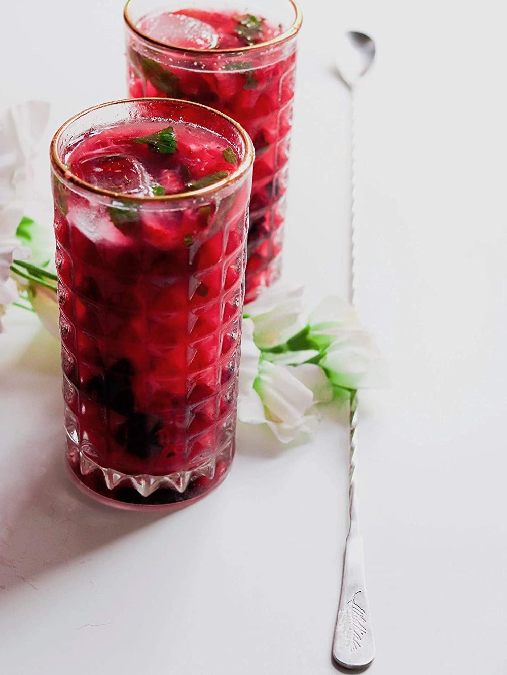 Two glasses of red berry iced tea on a white surface with flowers and a spoon.