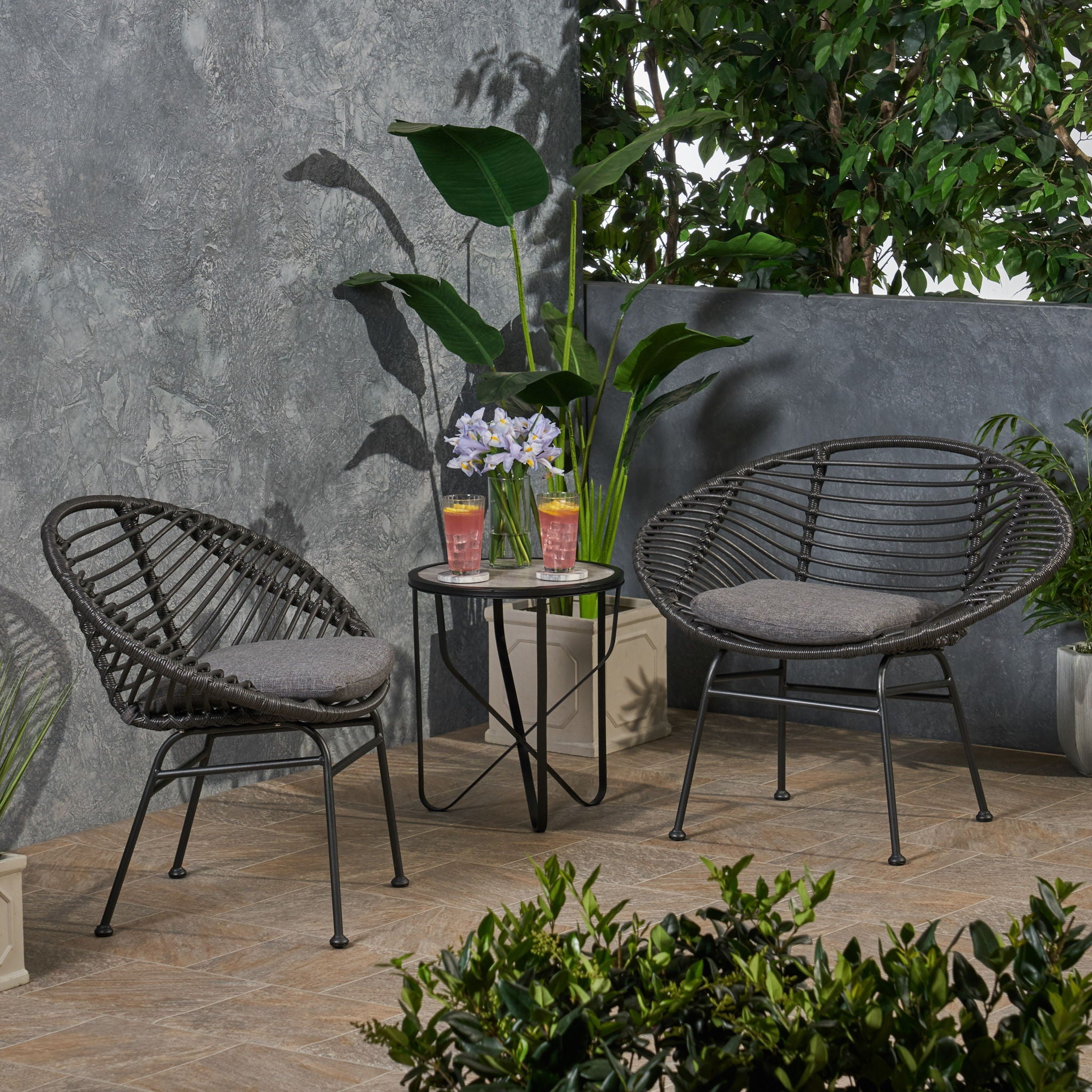 Two wicker chairs with a small table on a patio with plants and a gray wall.