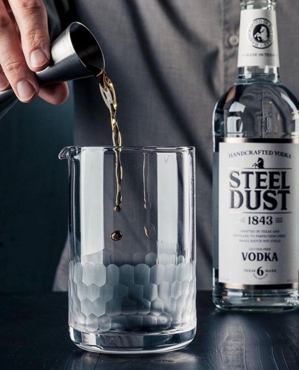 Person pouring a drink into a cocktail mixing glass with a bottle of Steel Dust Vodka in the background.