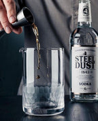 Person pouring a drink into a cocktail mixing glass with a bottle of Steel Dust Vodka in the background.