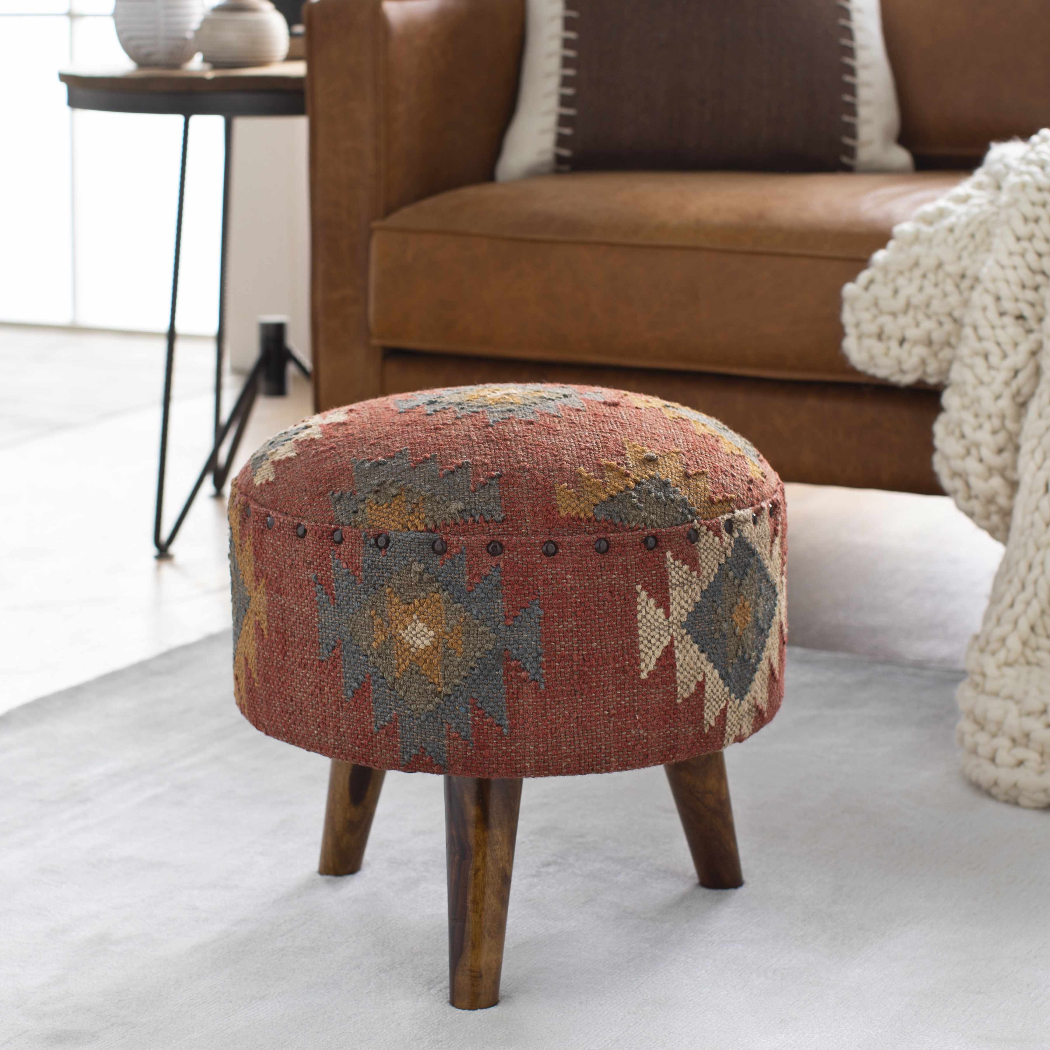 A round upholstered ottoman stool with a distressed geometric kilim pattern in rust red, beige, and gray, supported by four wooden legs. It is styled in a living room setting on a light gray rug in front of a brown leather sofa.