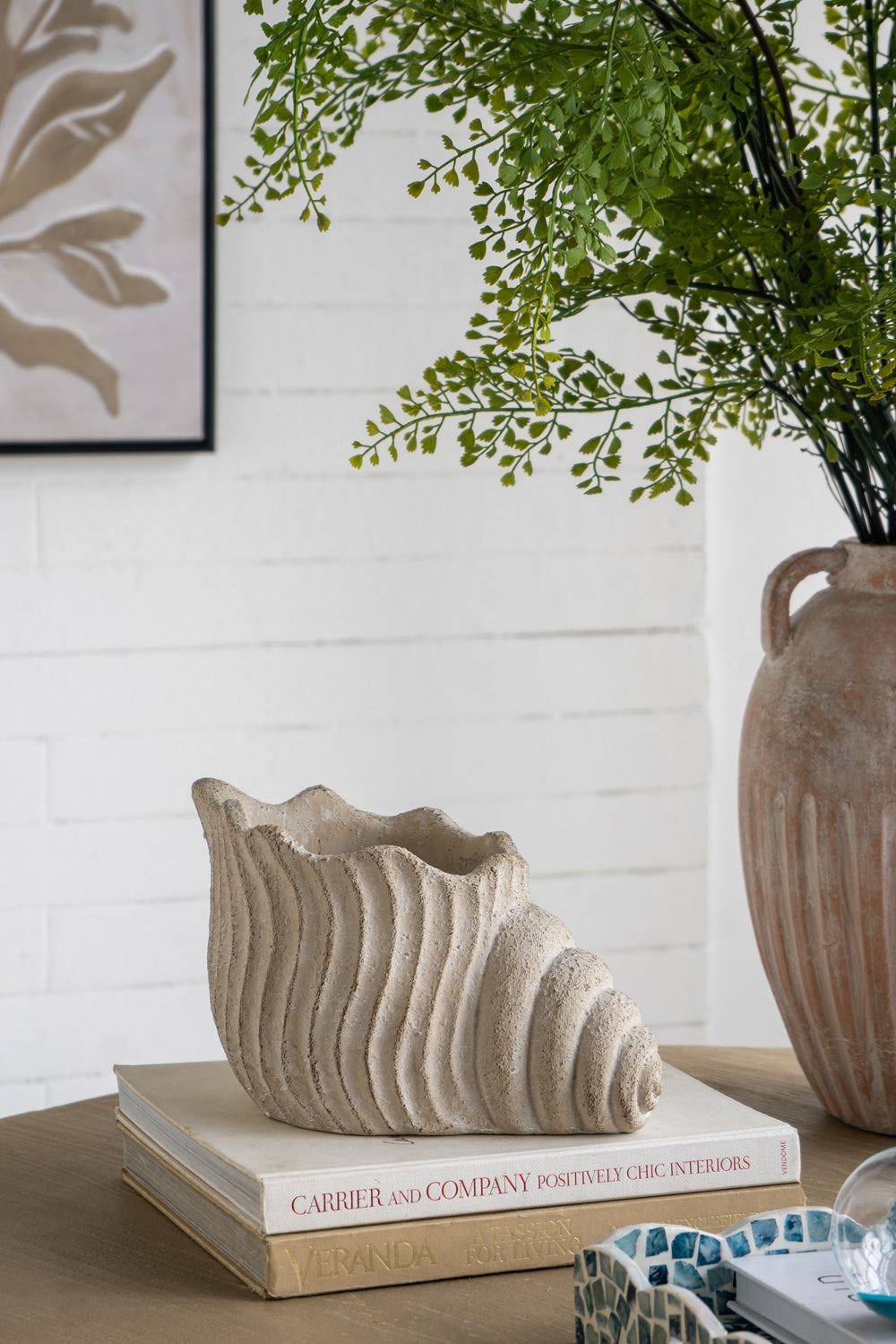 Decorative setting with a textured vase on books, a plant, and a framed picture on a white brick wall.