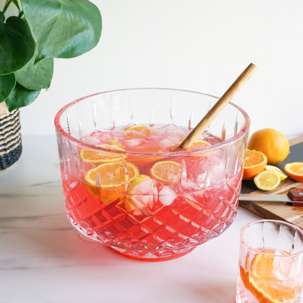 Clear glass punch bowl with pink liquid, oranges, and lemons on a white surface with a plant in the background.