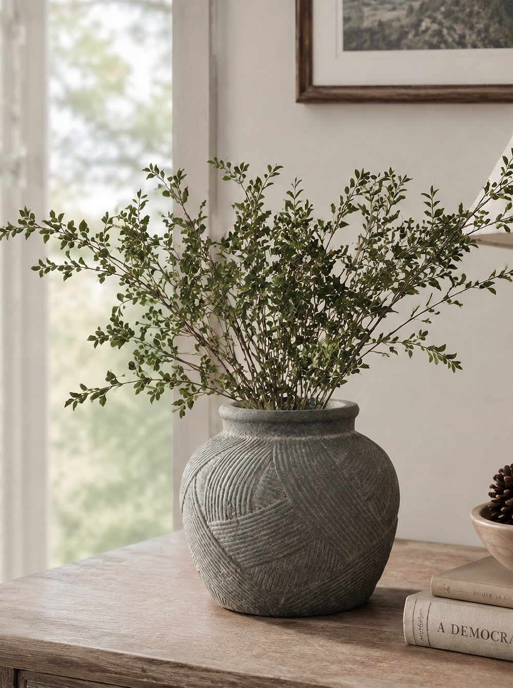 A textured gray ceramic pot with carved linear patterns, filled with green leafy branches, displayed on a light wood table in a softly lit room.