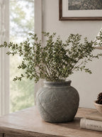 A textured gray ceramic pot with carved linear patterns, filled with green leafy branches, displayed on a light wood table in a softly lit room.