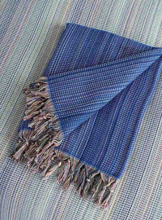 Folded dark blue king size blanket with multicolor fringe on a woven surface