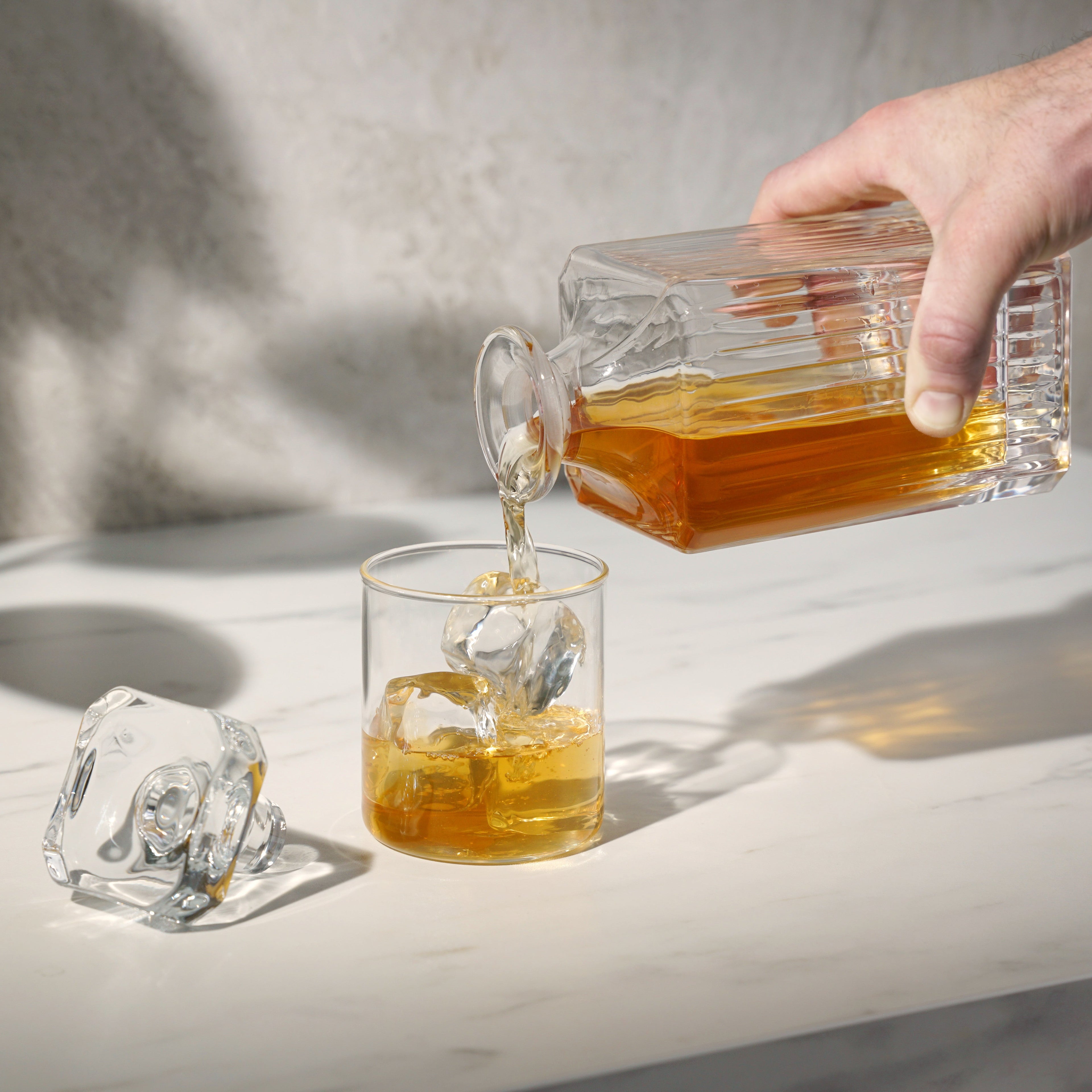 Hand pouring amber liquid from a glass container into a glass with ice cubes on a light surface.