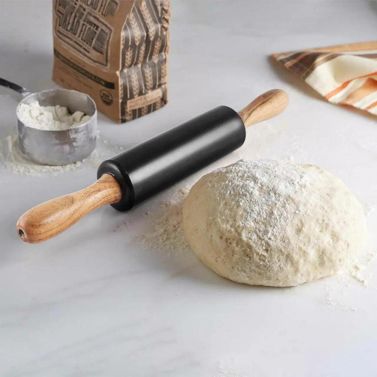 A black rolling pin with light wood handles lies on a white marble-like surface beside a flour-dusted ball of dough, a flour sack, a measuring cup, and a striped cloth.