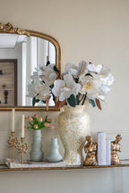 A styled interior scene showing a large textured cream vase filled with white magnolia flowers on a mirrored console shelf, alongside smaller vases, candles, books, and gold-toned figurines against a warm beige wall.