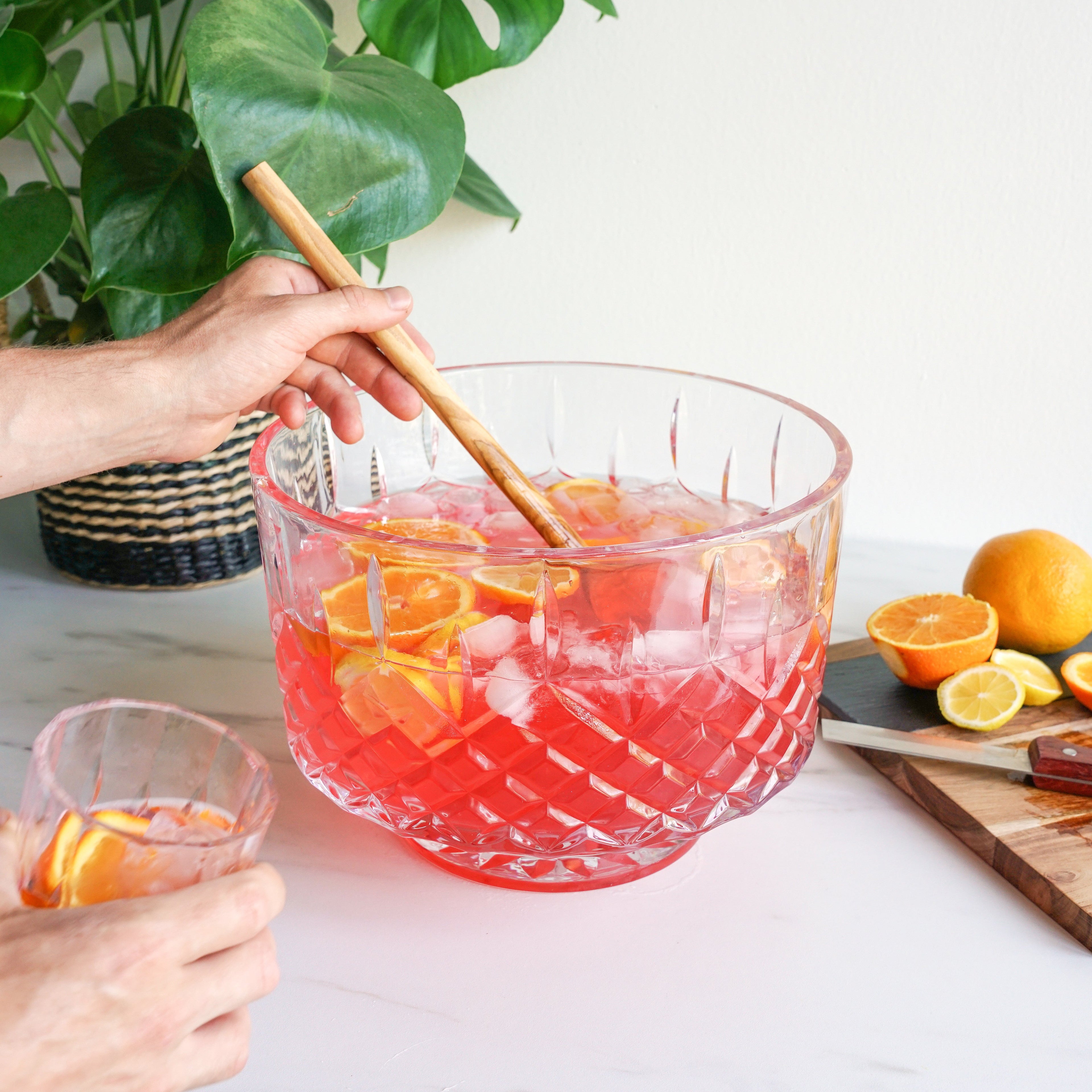 Person stirring a large glass bowl of pink lemonade with oranges and ice, surrounded by lemons and oranges on a white surface.