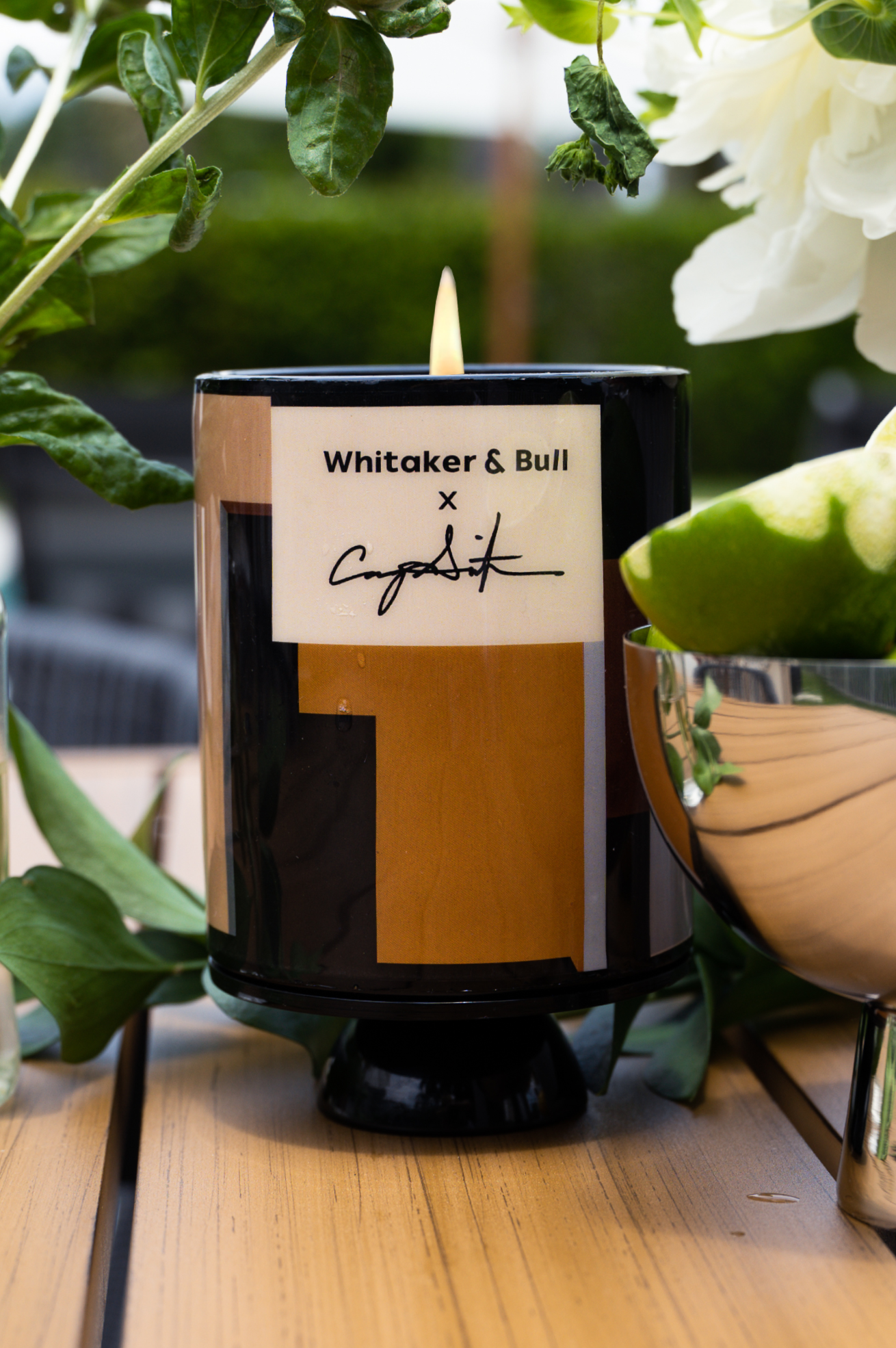 Candle with a label on a wooden surface surrounded by greenery