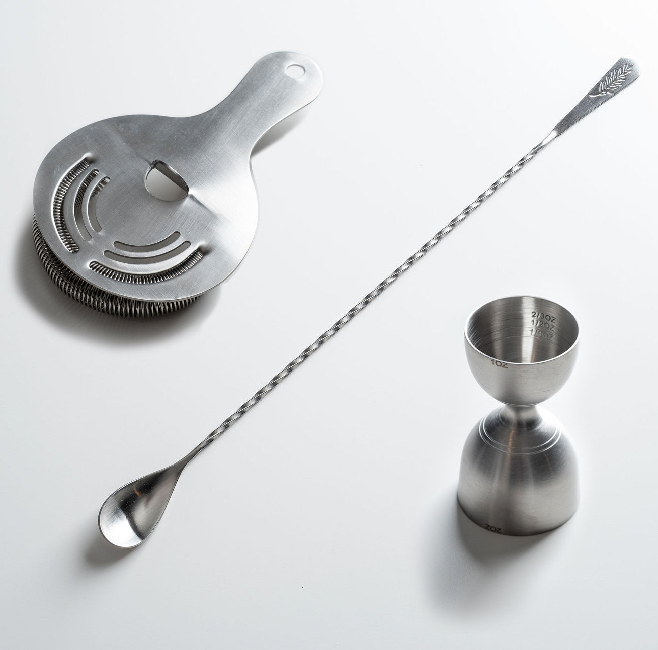 Set of metal cocktail tools including a strainer, jigger, and long spoon on a white background